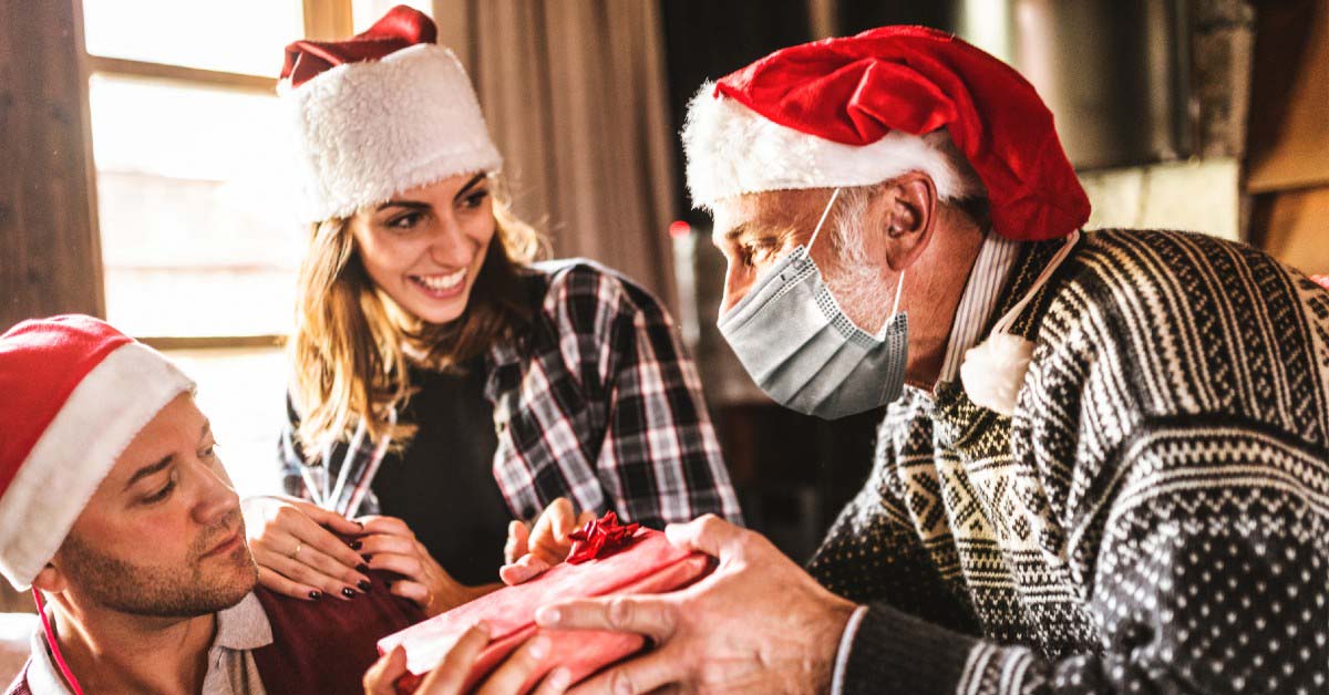 Older man wearing medical face mask gives a holiday gift to younger man wearing no mask as younger woman with no mask watches. All are wearing Santa hats.