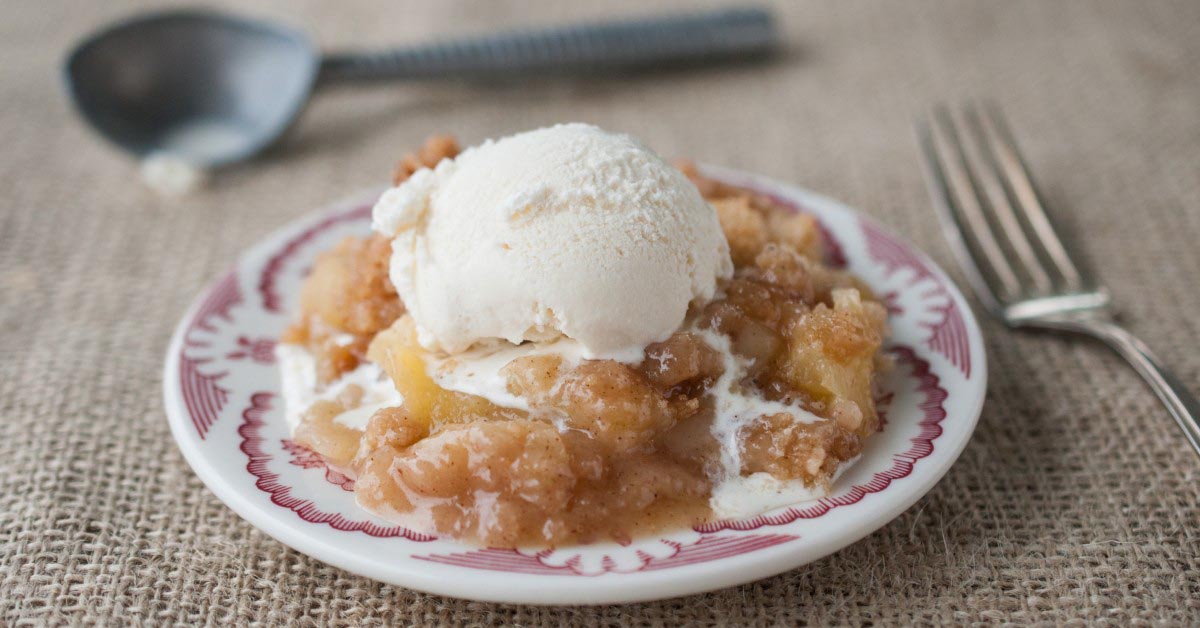 A serving of Apple Crisp on a small plate with melted vanilla ice cream on top.